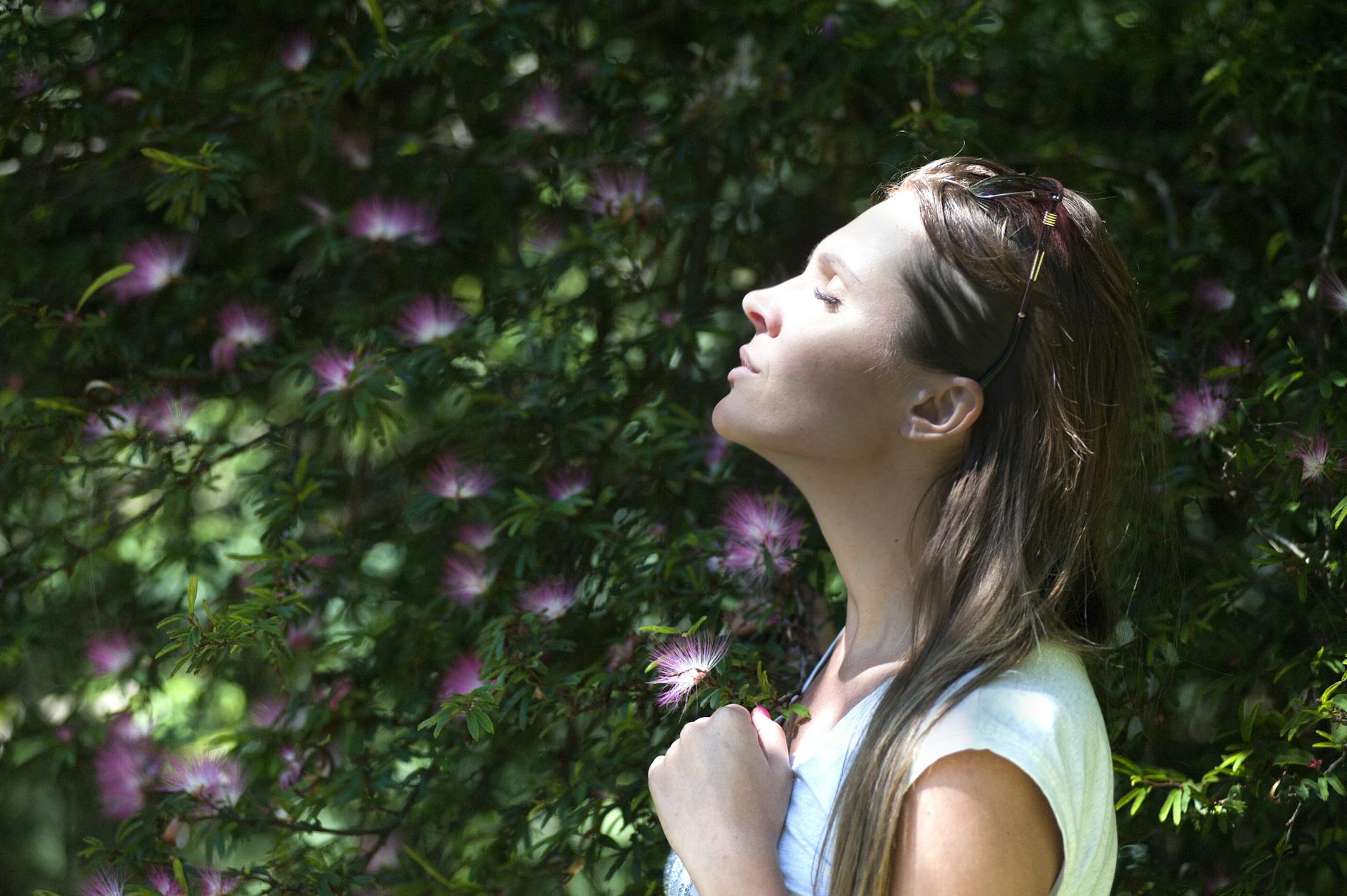 woman closing her eyes against sun light standing near purple petaled flower plant. anxiety calm
